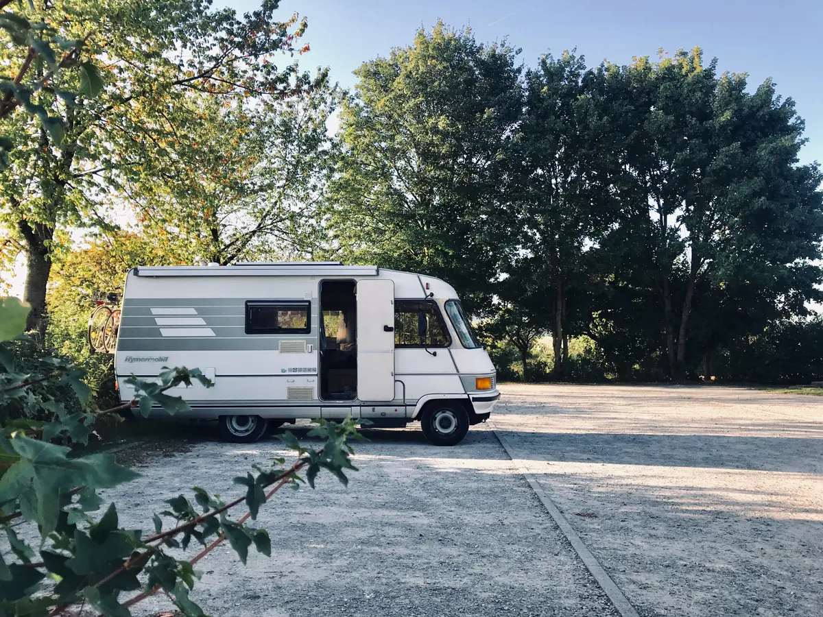 A classic Hymer campervan parked in an empty carpark surrounded by trees on a sunny day