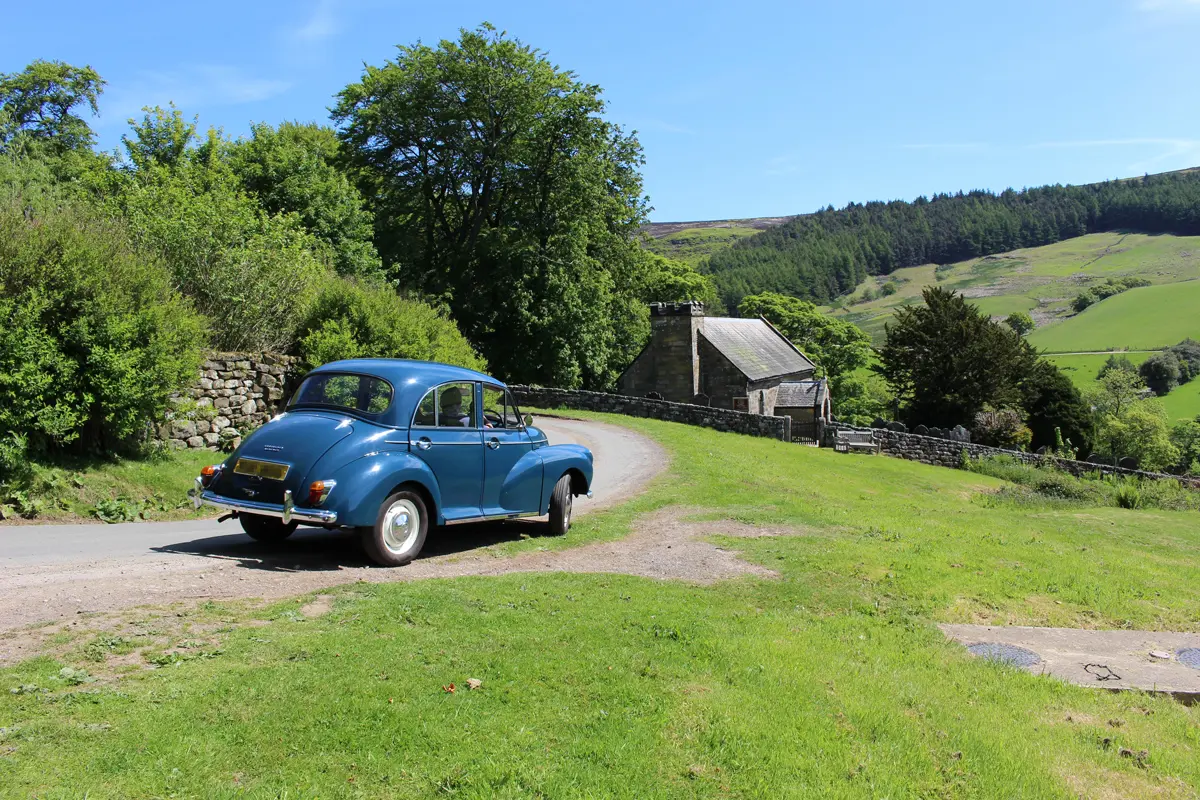 A Morris Minor driving along a thin country road next to a farm