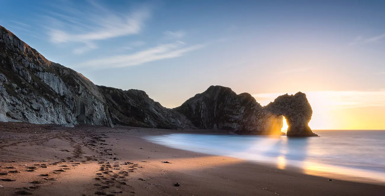 Durdle Door beach with the rising sun shining through the arch in the cliff