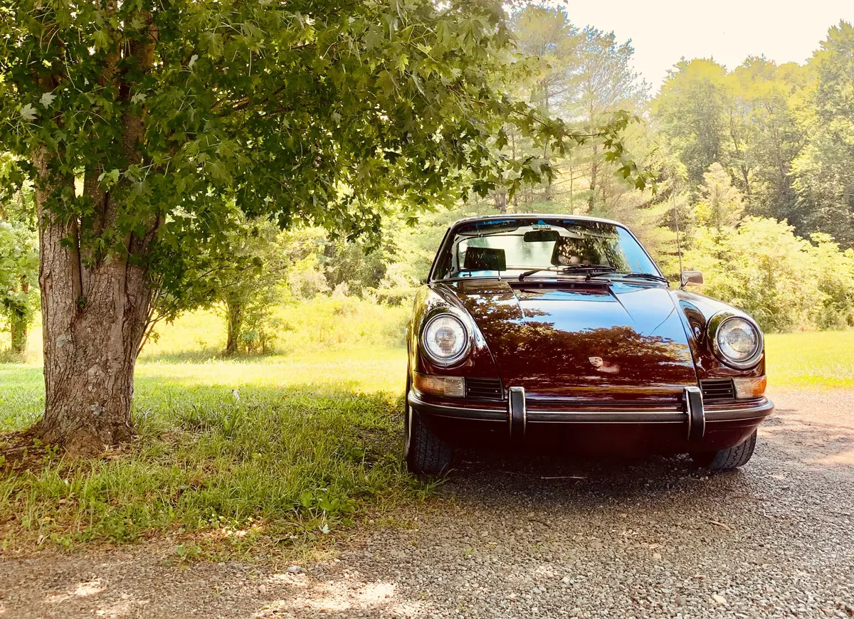 A burgundy classic Porsche parked next to a tree on a gravel road