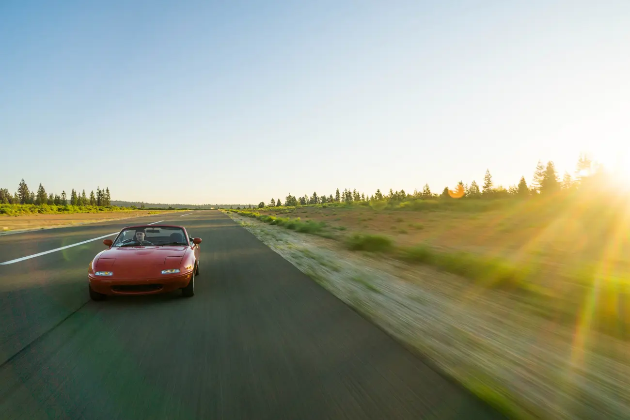 A classic Mazda driving along an open road with the sun setting in the distance