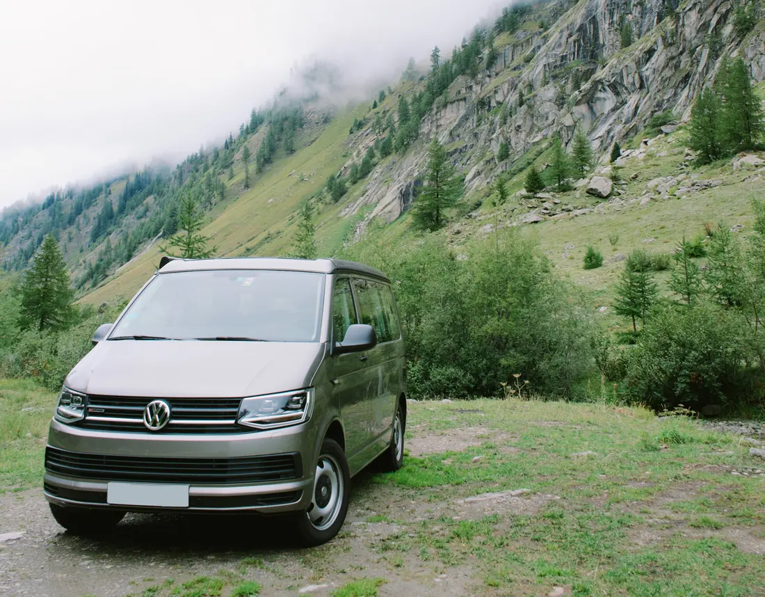 A brown Volkswagen T6 Transporter parked on a grassy hillside in an alpine region
