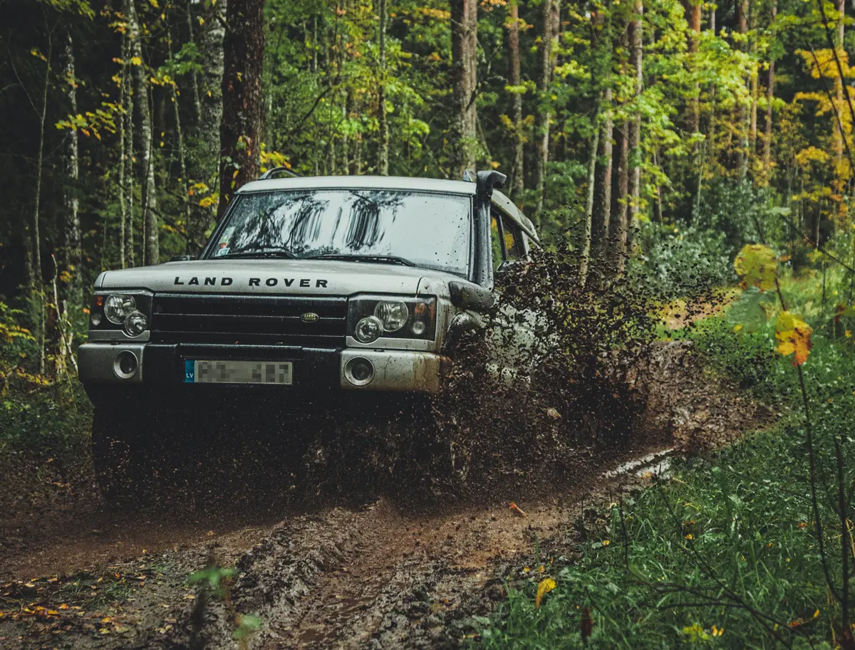A classic Land Rover Discovery driving along a dirt track in a wooded area with mud spraying from its wheels