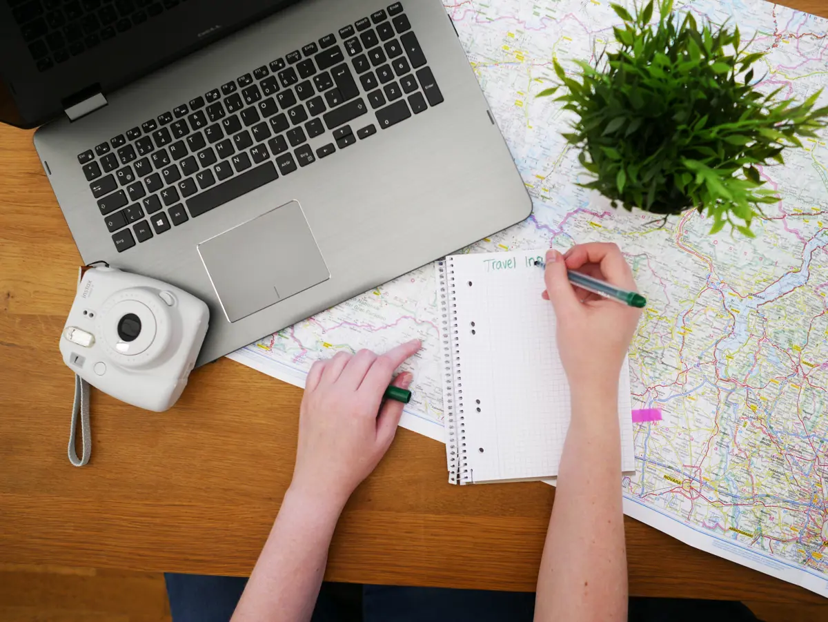 A person writing a travel plan on a notepad next to a laptop, a Polaroid camera and a map