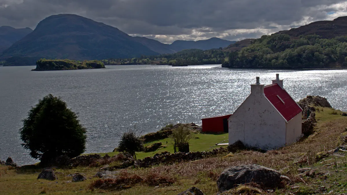 A white house on the edge of a loch with rolling hills in the distance