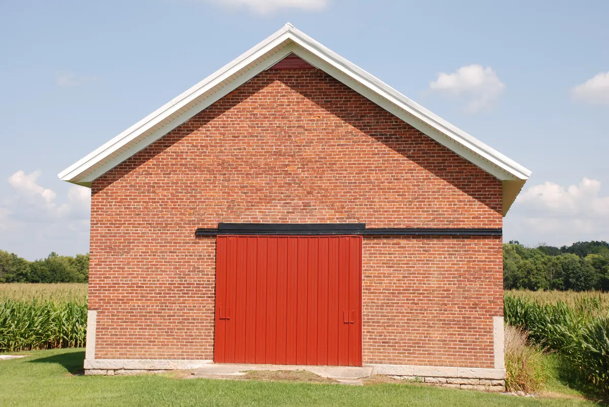 A large brick garage with a big red door on the edge of a crop field