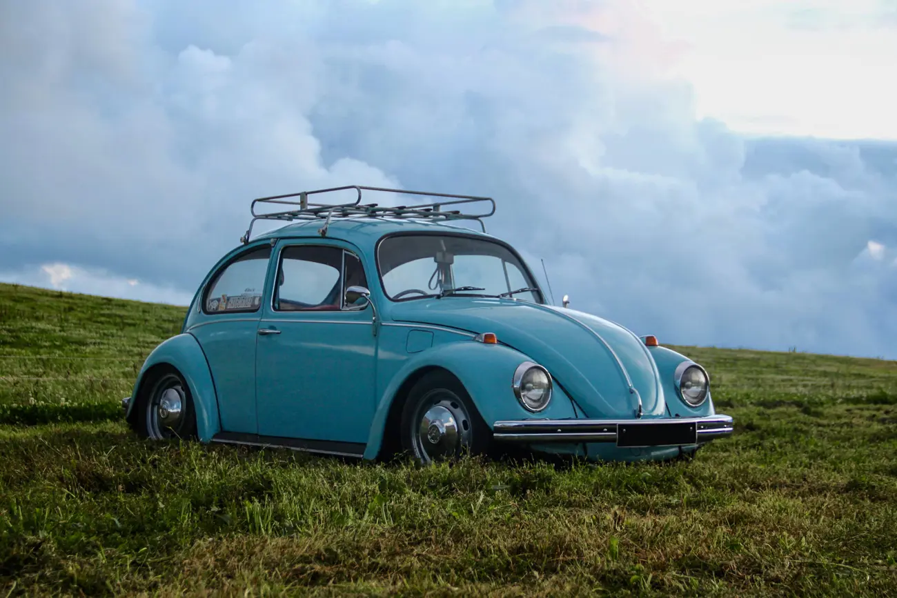 A blue classic VW Beetle with a roof rack parked on a grassy hill against a cloudy sky
