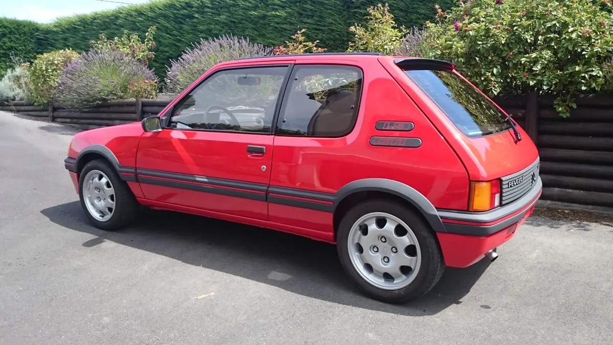 A rd Peugeot 205 GTi parked on a driveway next to flower beds on a sunny day