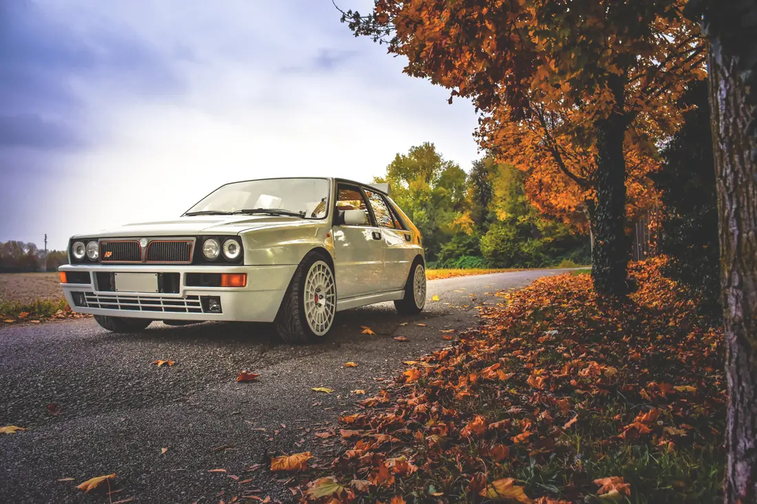 A white Lancia Delta driving on a country road with brown leaved trees along the side of the road