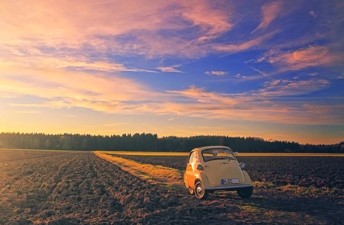 An Isetta bubble car parked on a grass track between ploughed fields at sunset