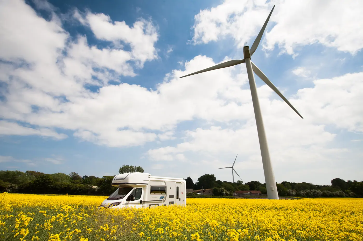 A motorhome driving on a road hidden by tall yellow crops and a wind turbine in the background