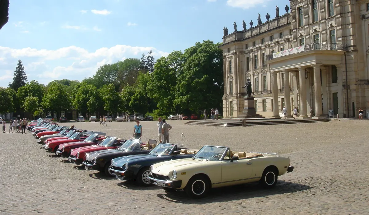 A car club of Fiat 124 owners parked in a line in-front of a historic building