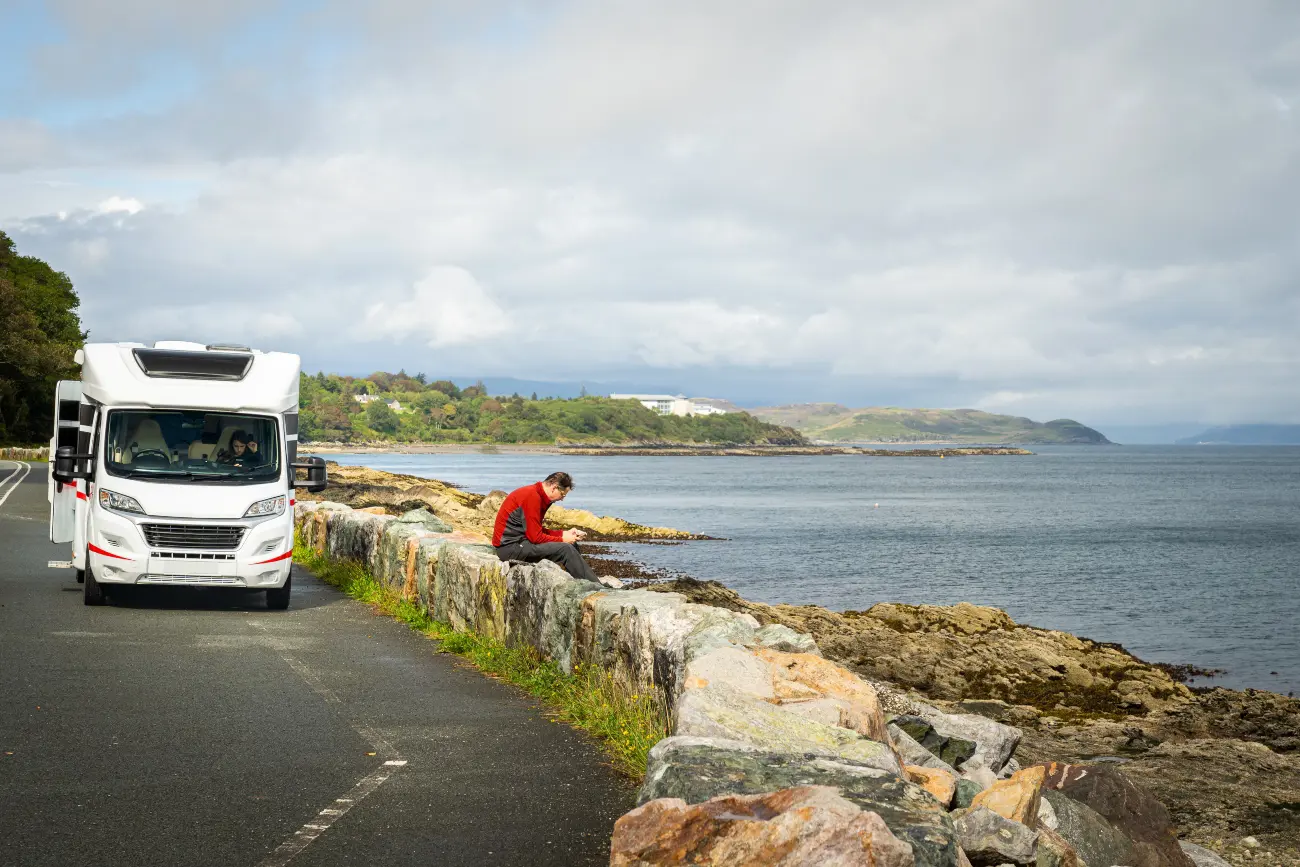 A parked campervan on the side of road with a man sat on a wall looking out at the river