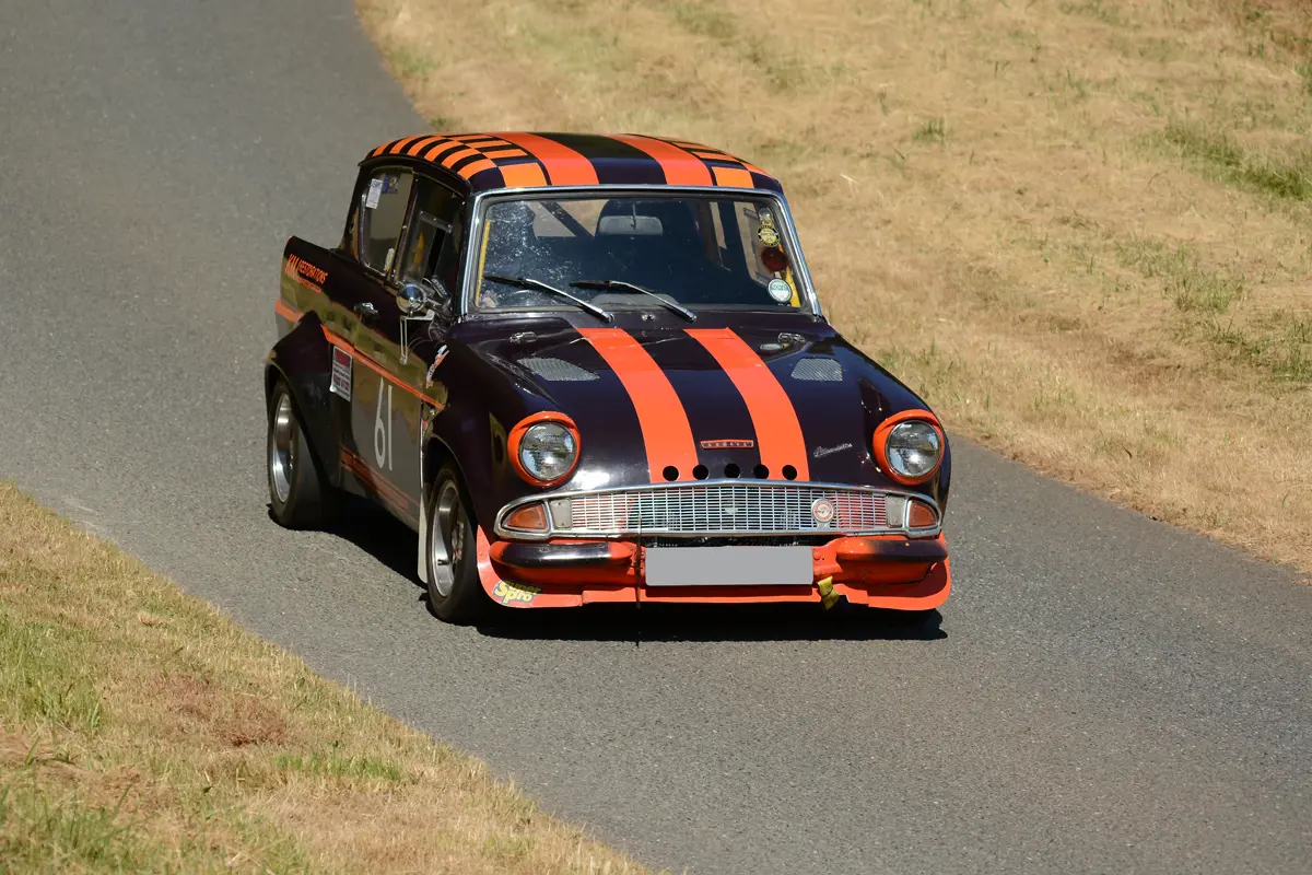 A Ford Anglia with racing stripes driving along a thin road in a field